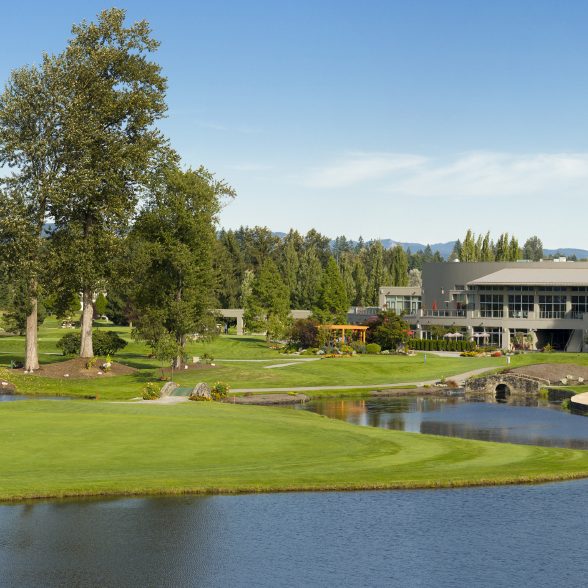 Landschaft mit Golfplatz, Wasser, Bäumen und einem modernen Clubhaus im Hintergrund.