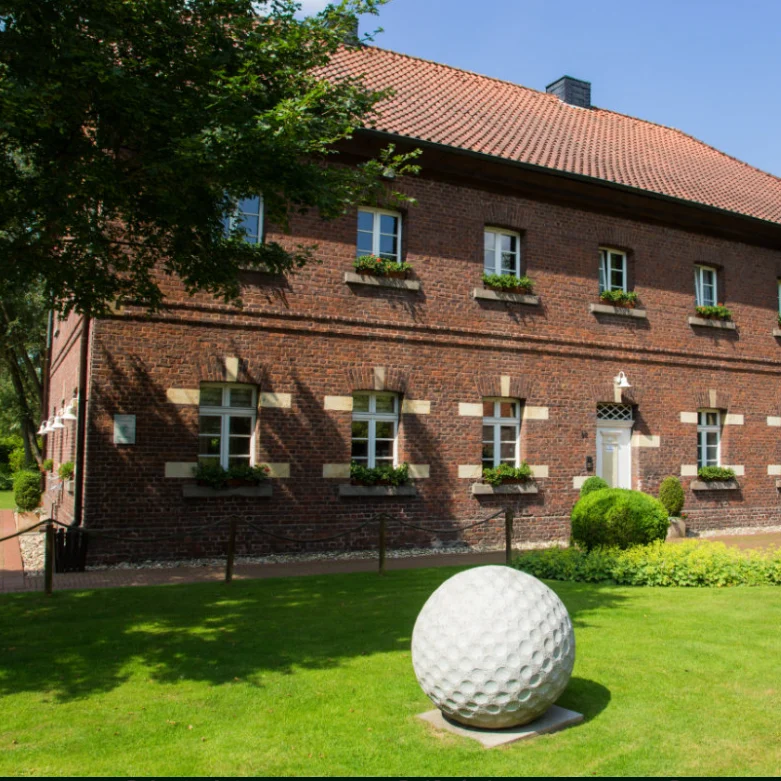 Rotes Backsteingebäude des Golfclubs Haus Leythe, neben einer großen weißen Golfball-Skulptur.