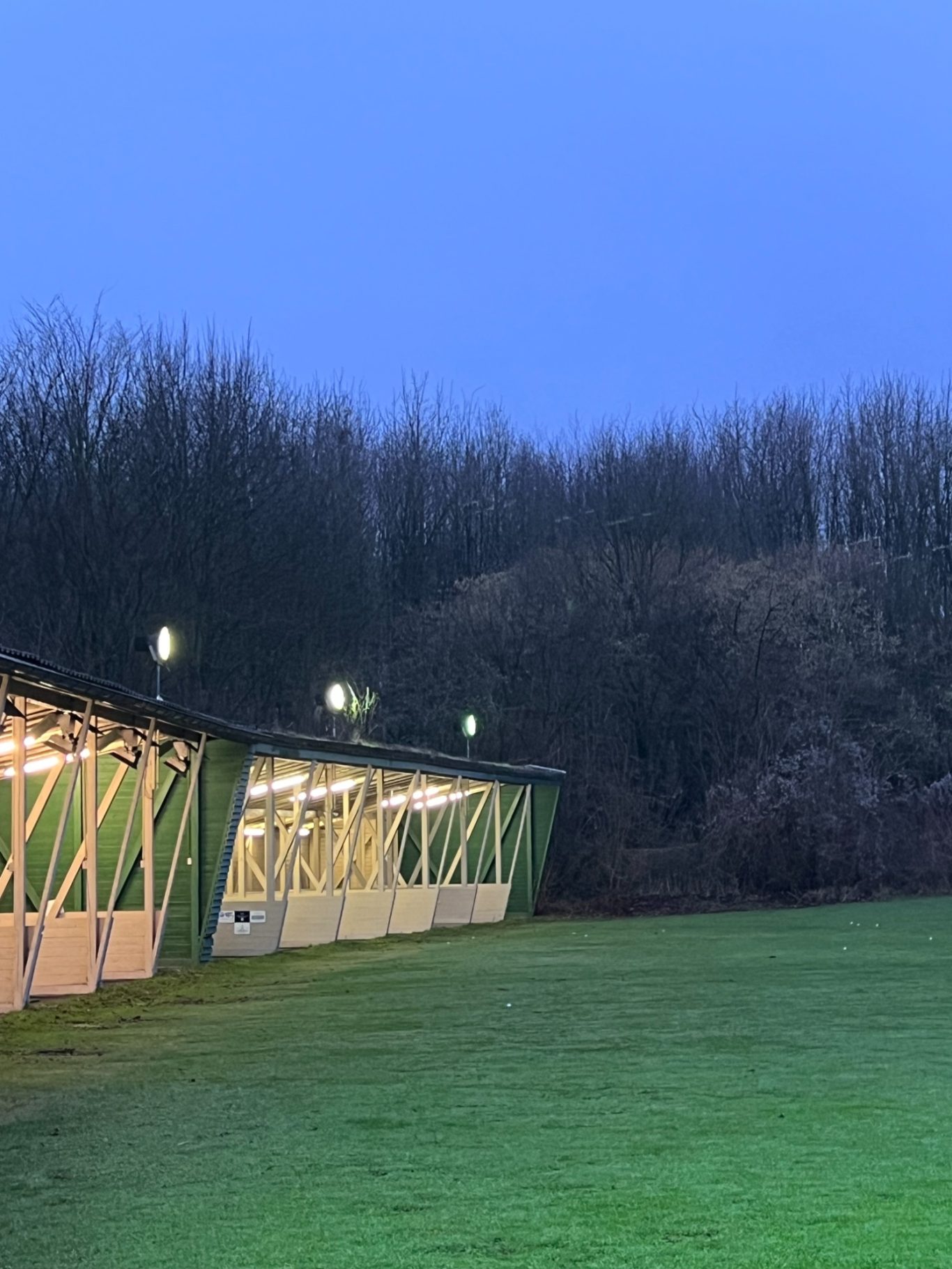 Driving Range mit mehreren beleuchteten Abschlagboxen unter einem dunkelblauen Himmel und Bäumen im Hintergrund.