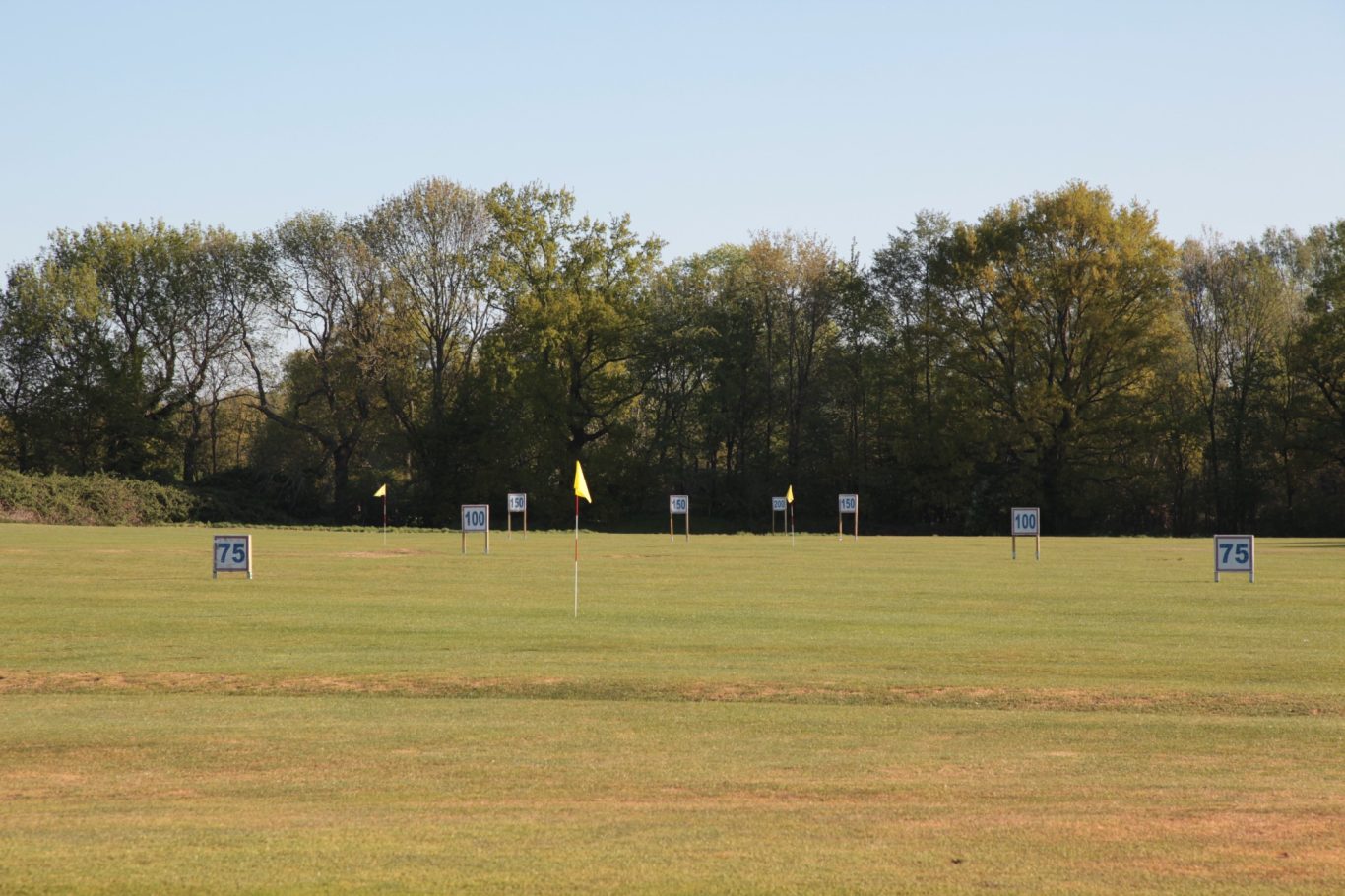 Driving Range mit Zielscheiben, Fahnen und Entfernungsangaben im Hintergrund unter blauem Himmel.